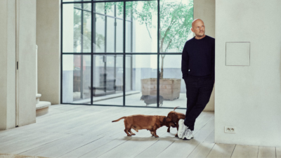 alt="Vincent Van Duysen we wnętrzu utrzymanym w minimalistycznym stylu, z dużymi przeszkleniami i dwoma jamnikami, ukazującymi osobisty wymiar jego architektury."