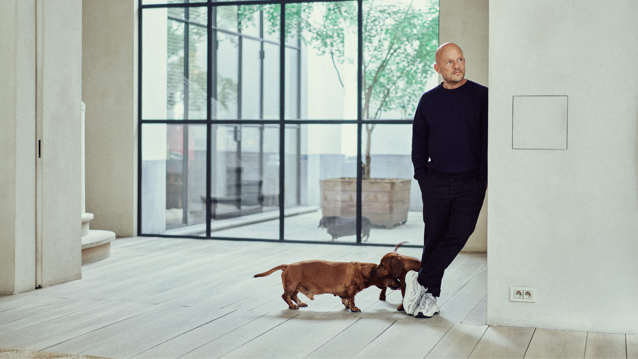 alt="Vincent Van Duysen we wnętrzu utrzymanym w minimalistycznym stylu, z dużymi przeszkleniami i dwoma jamnikami, ukazującymi osobisty wymiar jego architektury."